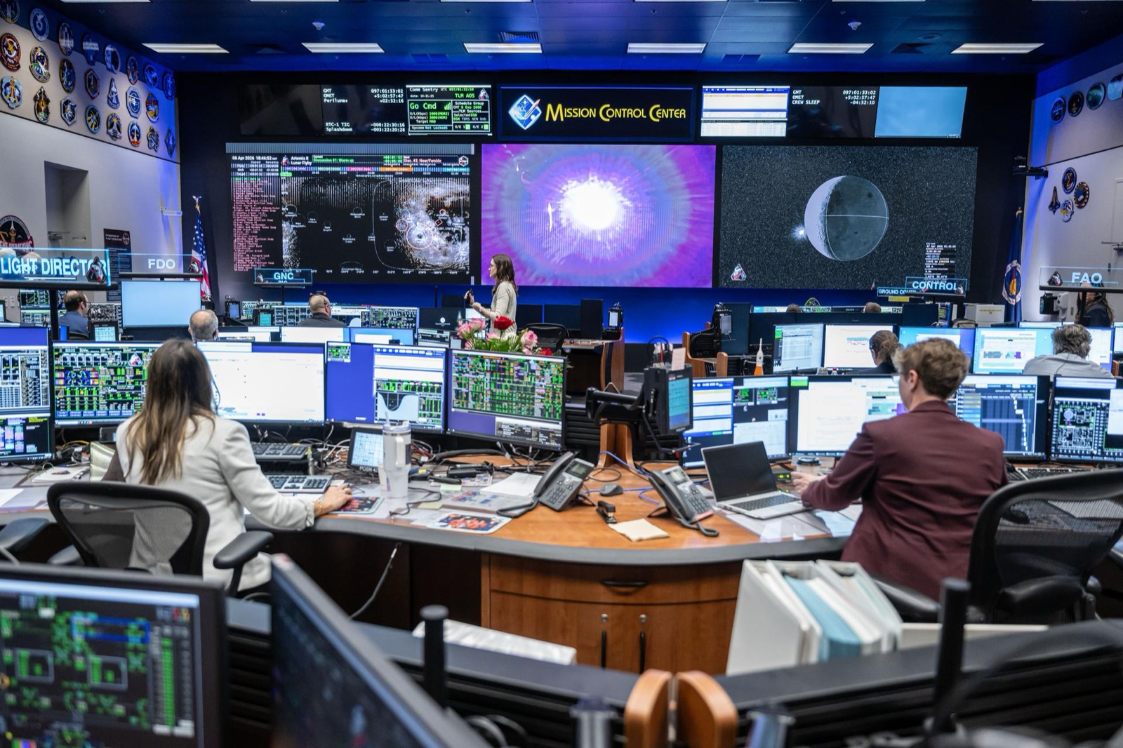 Flight controllers at their consoles in mission control during a crewed lunar flyby, the Moon on the main screen